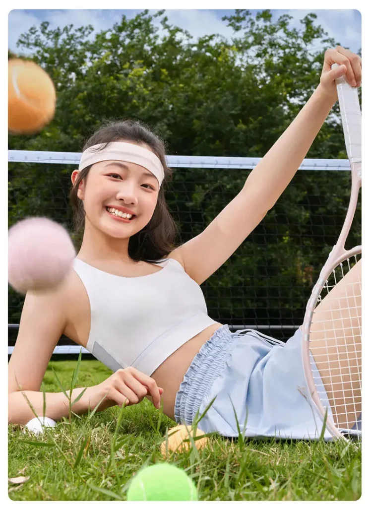Happy girl sitting on grass wearing a white Suyiyi seamless sports bra and light blue shorts, surrounded by tennis balls and holding a racket, emphasizing comfort and style in Suyiyi training bras