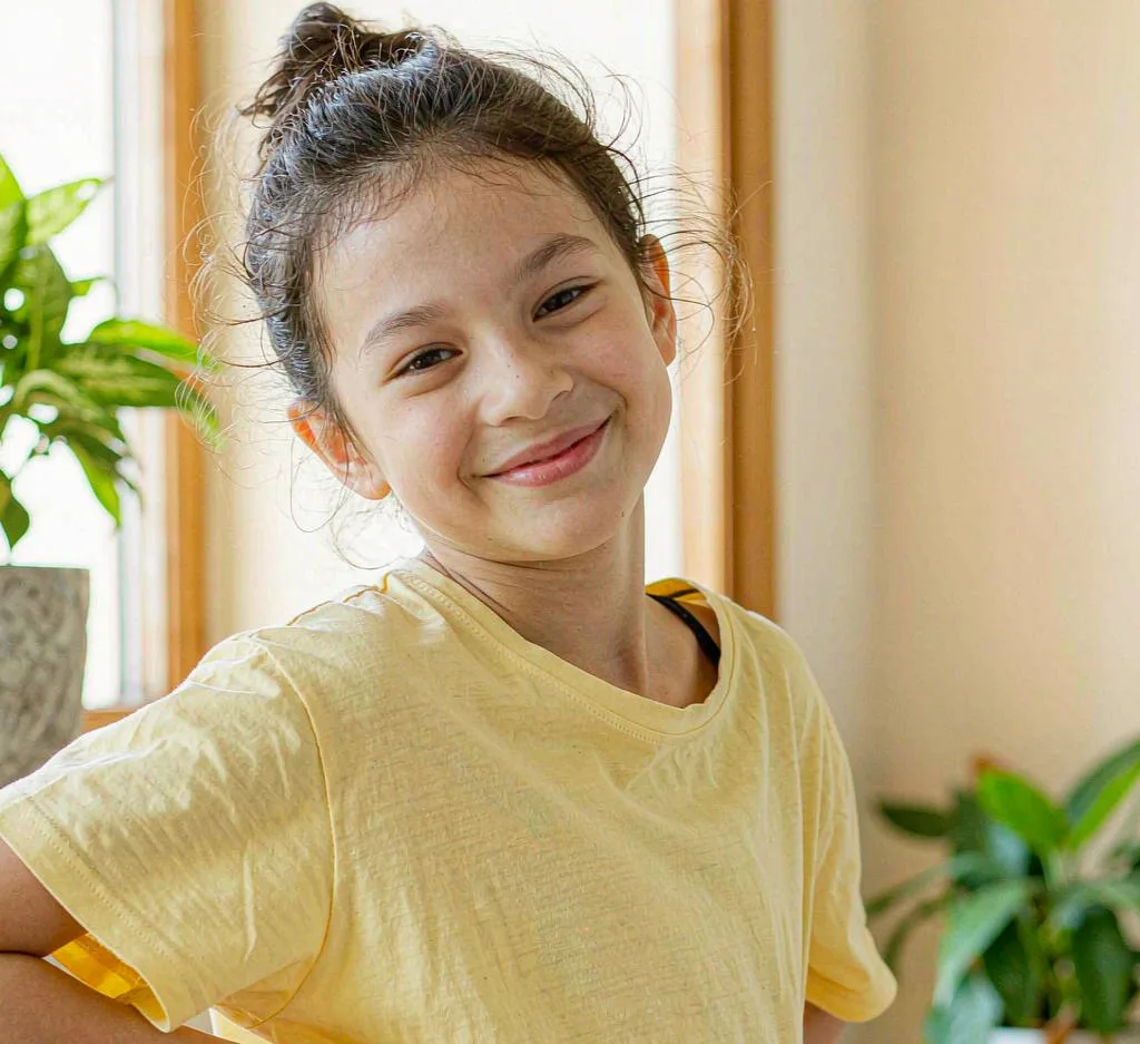 A tween girl smiles confidently while wearing a soft yellow t-shirt in a sunlit room.