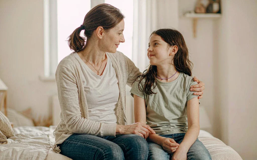 Mom talking calmly with her 10-year-old daughter on the edge of a bed — a reassuring moment about normal breast development and breast lumps in tweens
